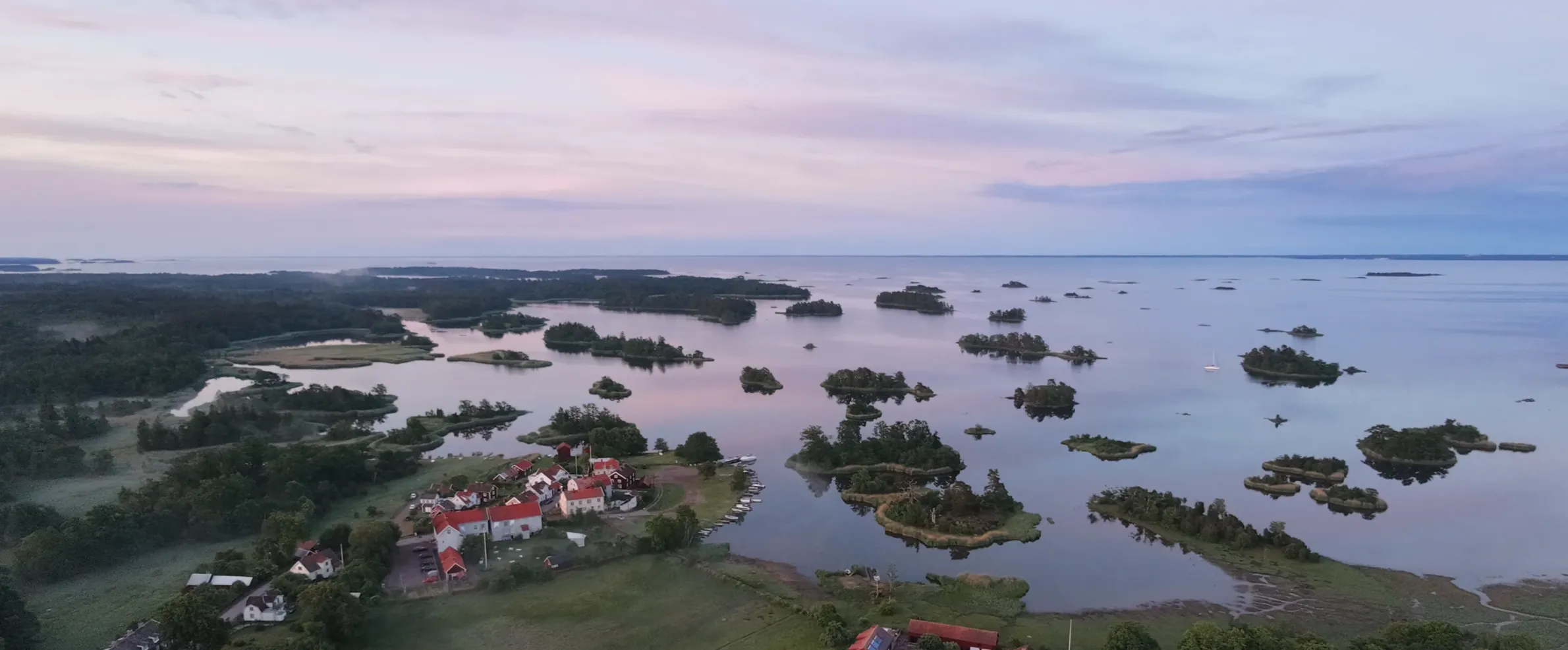 Flygvy över Pataholm — köpingens låga trähusbebyggelse vid vattnet, med skärgårdens holmar och grunda vikar ut mot en rosaskimrande horisont.